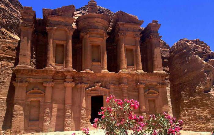 Jordanien | Kloster Ad Deir in Petra fotografiert aus der Nähe. Im Vordergund pinker Rhododendron vor den mächtigen roten Säulen des Eingangs zum Kloster
