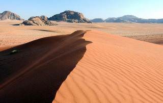 Jordanien | Eine von vielen Sanddünen im Wadi Rum. Ein kleiner Hügel aus rotem Wüstensand im Hintergrund eine Bergkette bei strahlend blauem Himmel