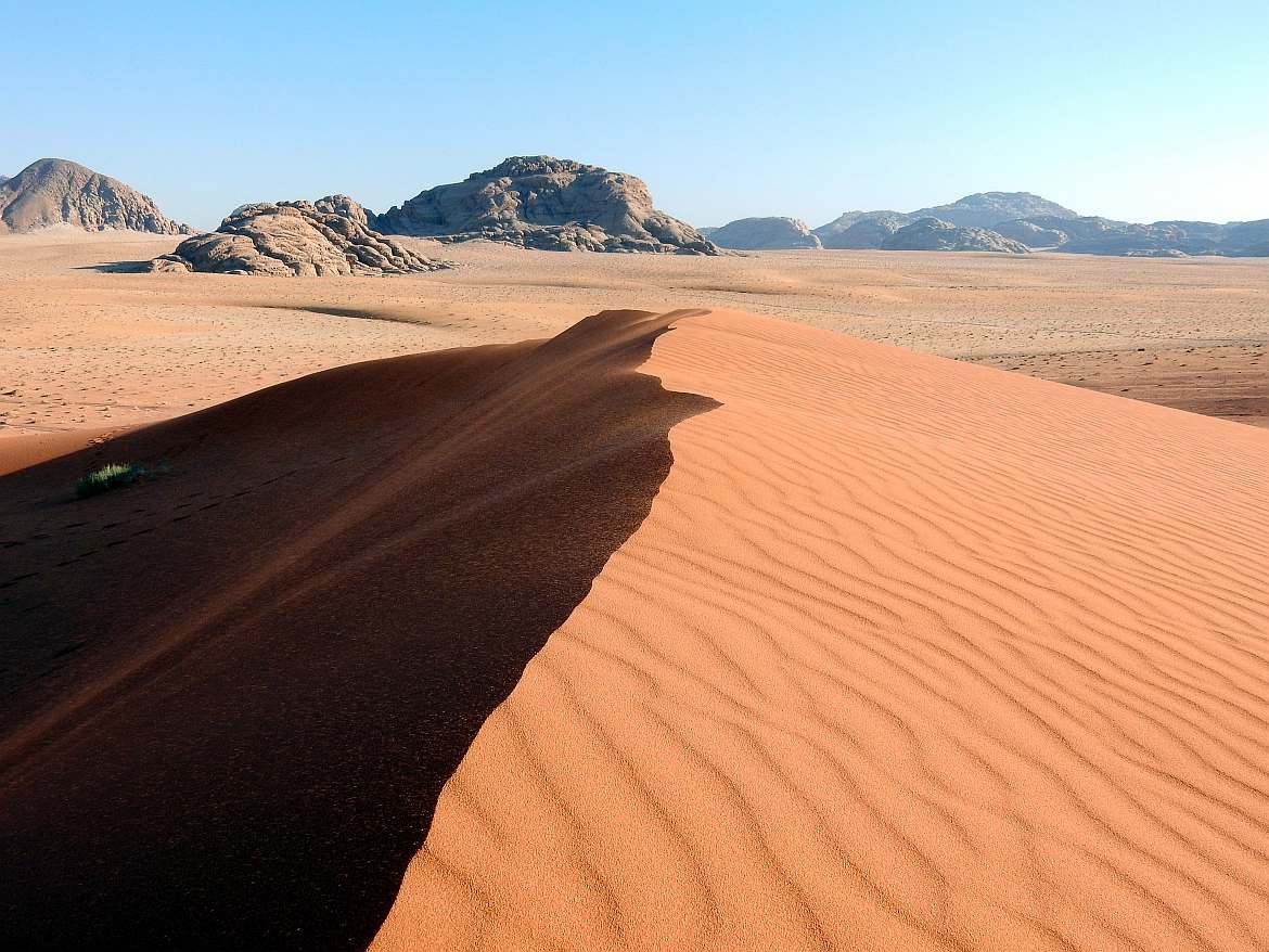 Jordanien | Eine von vielen Sanddünen im Wadi Rum. Ein kleiner Hügel aus rotem Wüstensand im Hintergrund eine Bergkette bei strahlend blauem Himmel