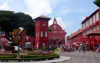 Malaysia | Zentrum der Stadt, der Roter Platz in Melaka. Rote Häuser und die Melaka Kirche aus der Kolonialzeit, Touristen und eine runde Blumeninsel mit Wegweisern in verschiedene Länder. Eine der Top-Sehenswürdigkeiten