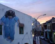 Malaysia | An die Hauswand gemalte Straßenkunst in George Town, Penang bei Sonnenuntergang. Ein Mädchen im blauben Overall mit schwarzen Zöpfen stützt sich auf ihren Händen auf zwei Dächer von Fenstern