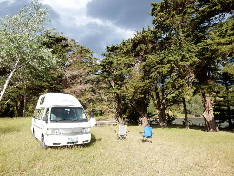 Neuseeland | Nordinsel, Simpsons Beach Campingplatz bei Whitianga in der Coromandel. Unser Camper und zwei Klappstühle stehen auf der grünen Wiese mit Blick durch Bäume auf den Strand mit schwarzen Wolken am Himmel und trotzdem Sonnenschein