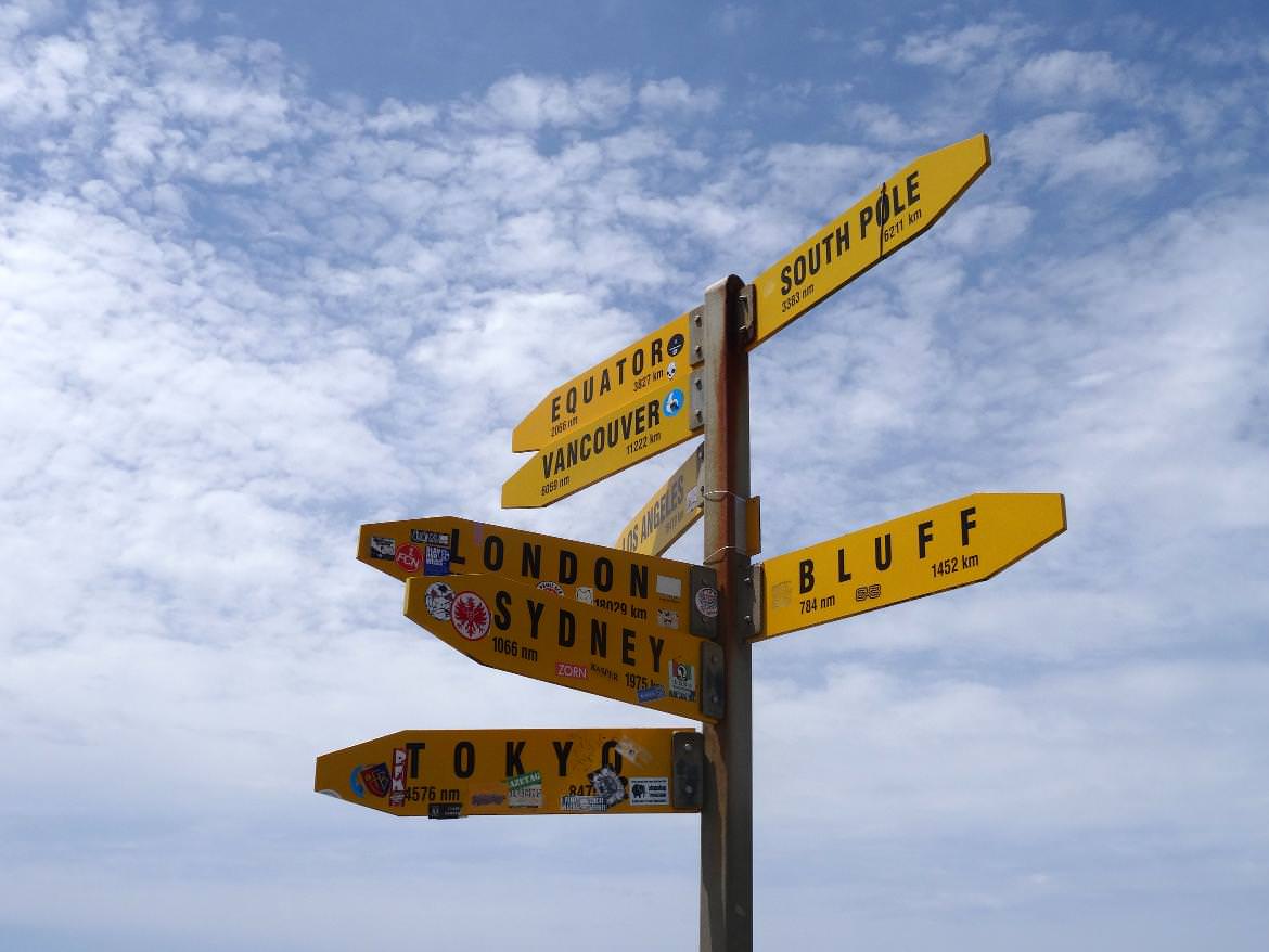 Neuseeland | Nordinsel, Hoher Norden, Weltweite Entfernungen vom Cape Reinga in Neuseeland. Gelbe Schilder in schwarzer Aufschrift vor blau-weißem Himmel. Tipps zur Entscheidung im Detail findest Du im Artikel
