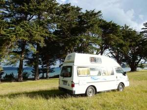Neuseeland | Nordinsel, Unser weißer Wendekreisen High-Top-Camper am Simpsons Beach Campingplatz in Whitianga. Stellplatz mitten in einer grünen WIese mit Blick auf das türkisfarbene Meer und schwarzen Vulkansand