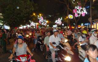 Vietnam | Süden, Mopedfahrer von Ho Chi Minh City. Schlangen von Mopeds mit mehreren Menschen auf einem Moped bei Nacht mit bunter Weihnachtsbeleuchtung in den Straßen. Saigon-Sehenswürdigkeiten, Tipps & Highlights findest Du in unserem Reisebericht