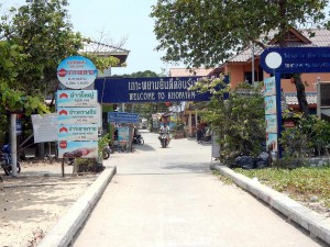 Thailand | Ankunft auf Ko Phayam. Ein dunkelblaues Schild mit weißer Aufschrift "Welcome to Ko Phayam" im Vordergrund, der Hauptort mit einigen Schildern und Shops im Hintergrund