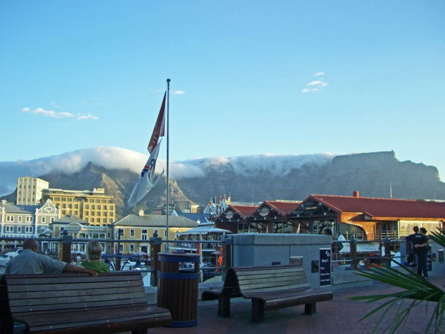 Südafrika | Kapstadt, V&A Waterfront mit Blick auf den Tafelberg samt Tischtuch bei blauem Himmel im Vordergrund ein Paar auf einer Bank von hinten fotografiert